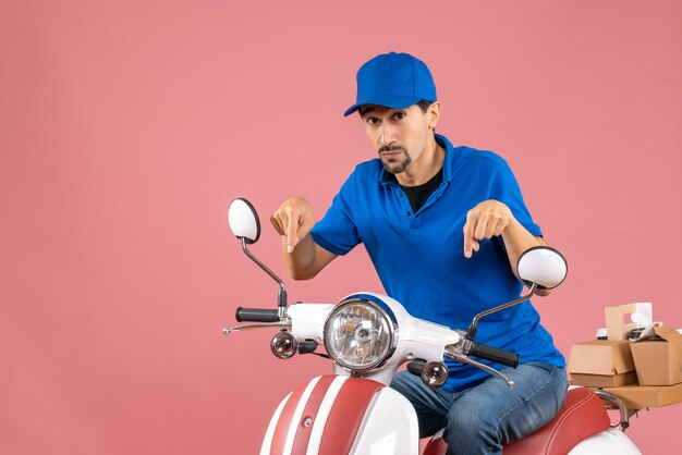 Vista frontal del tipo de mensajero con sombrero sentado en scooter y apuntando hacia abajo sobre fondo melocotón pastel