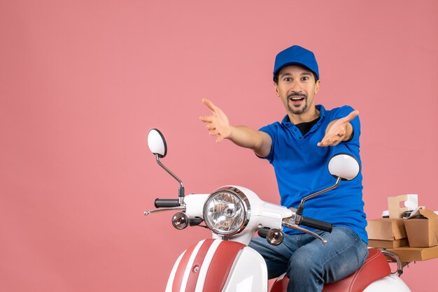 Vista frontal del tipo de mensajero con sombrero sentado en scooter recibiendo algo sobre fondo melocotón pastel