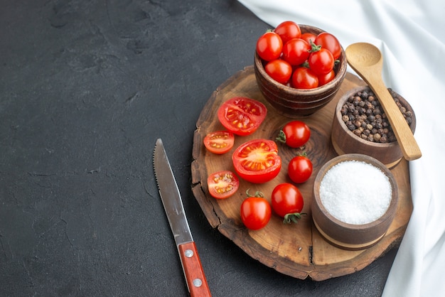 Vista frontal de todo el corte de tomates frescos y especias sobre tabla de madera, cuchillo de toalla blanca en el lado izquierdo sobre una superficie negra con espacio libre