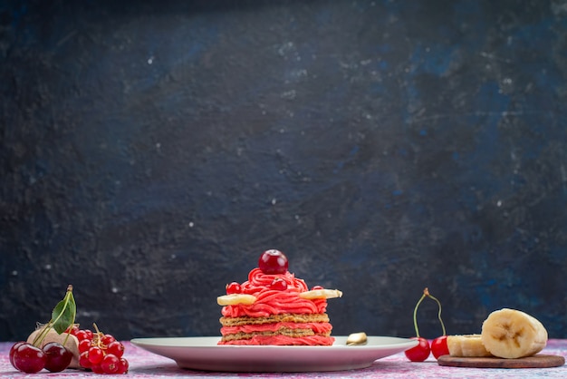 Vista frontal de la torta de crema roja dentro de la placa blanca con frutas frescas en la oscuridad