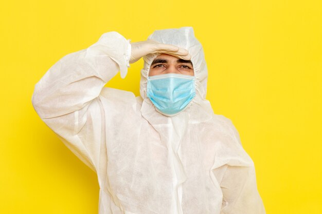 Vista frontal del trabajador científico masculino en traje blanco de protección especial con máscara estéril mirando a lo lejos en la pared amarilla