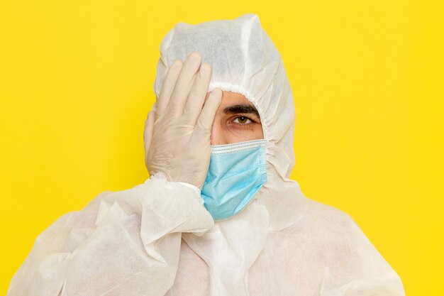 Vista frontal del trabajador científico masculino en traje blanco de protección especial con máscara estéril que cubre la mitad de su rostro en la pared amarilla