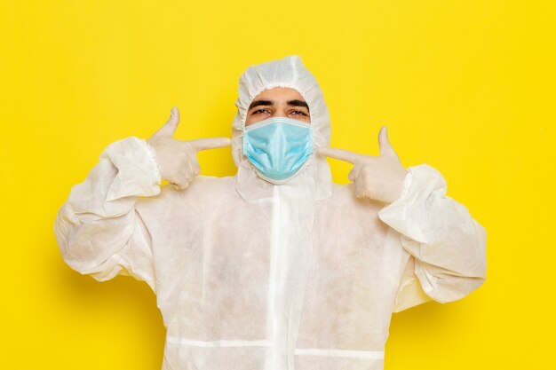 Vista frontal del trabajador científico masculino en traje blanco de protección especial con máscara en la pared amarilla