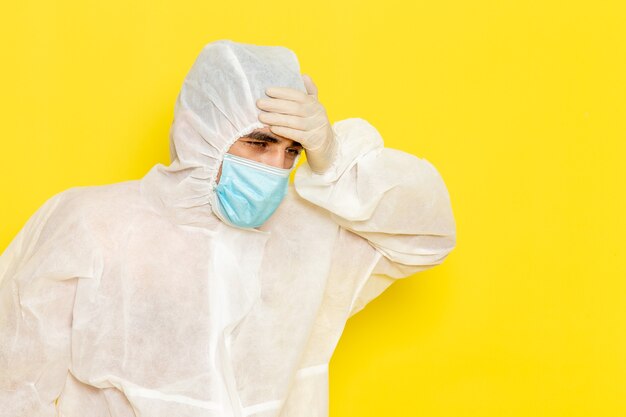 Vista frontal del trabajador científico masculino en traje blanco protector especial con máscara estéril que tiene dolor de cabeza en la pared amarilla