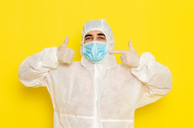 Vista frontal del trabajador científico masculino en traje blanco protector especial con máscara en la pared de color amarillo claro