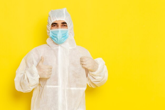 Vista frontal del trabajador científico masculino en traje de protección especial y con máscara posando en la pared de color amarillo claro