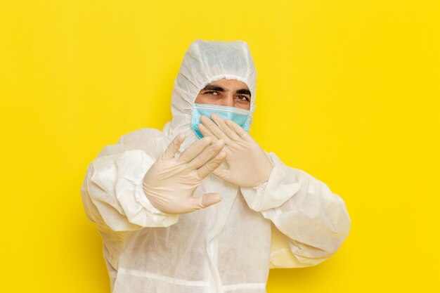 Vista frontal del trabajador científico masculino en traje de protección especial y con máscara posando en la pared de color amarillo claro