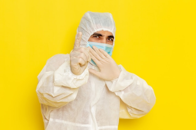 Vista frontal del trabajador científico masculino en traje de protección especial y con máscara que amenaza en la pared de color amarillo claro