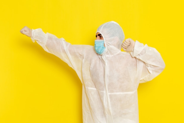 Vista frontal del trabajador científico masculino en traje de protección especial y con máscara simplemente posando en la pared de color amarillo claro