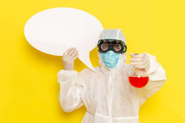 Vista frontal trabajador científico masculino en traje de protección especial con matraz con solución roja y letrero blanco en el escritorio amarillo