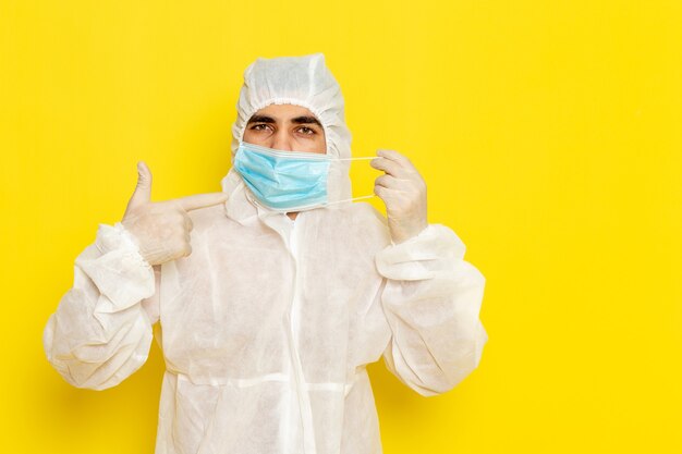 Vista frontal del trabajador científico masculino en traje de protección especial quitándose la máscara en la pared de color amarillo claro