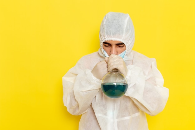 Vista frontal del trabajador científico masculino en traje de protección especial sosteniendo el matraz con solución que lo huele en la pared amarilla