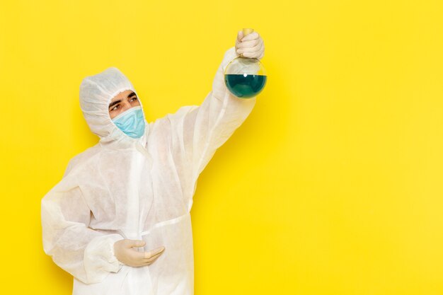 Vista frontal del trabajador científico masculino en traje de protección especial sosteniendo el matraz con solución verde en la pared amarilla