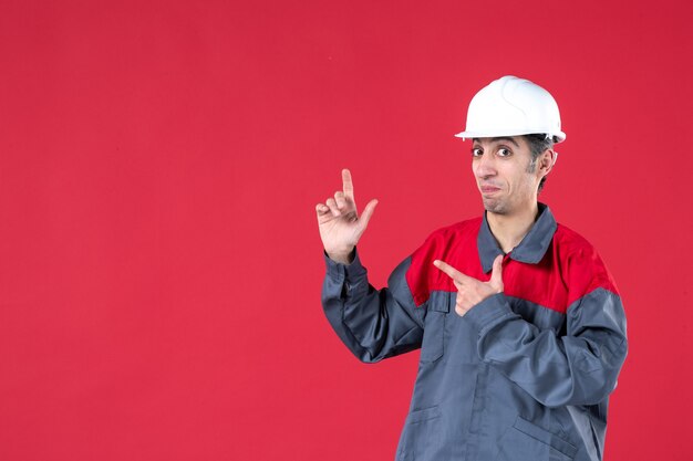 Vista frontal del trabajador joven confundido en uniforme con casco apuntando hacia arriba en la pared roja aislada