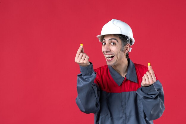 Vista frontal del trabajador joven feliz en uniforme con casco y sosteniendo tapones para los oídos en la pared roja aislada