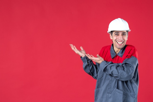 Vista frontal del trabajador joven sonriente en uniforme con casco y mostrando algo en el lado derecho en la pared roja aislada