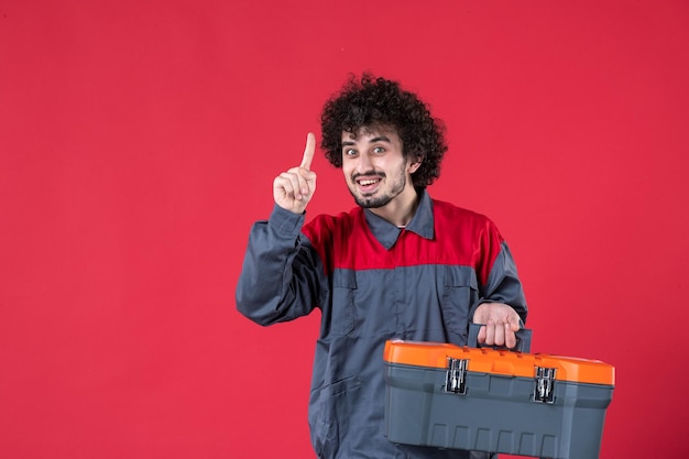 Vista frontal trabajador masculino con caja de herramientas sobre fondo rojo color de la foto del trabajador de la casa instrumento trabajo emoción uniforme mecánico