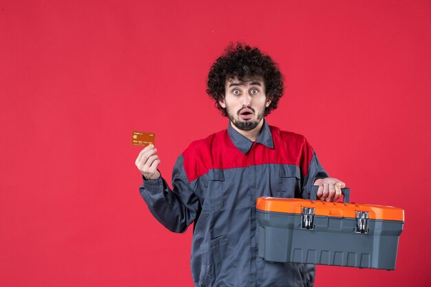 Vista frontal trabajador masculino con caja de herramientas y tarjeta bancaria sobre fondo rojo foto color trabajador casa uniforme de trabajo instrumentos mecánicos