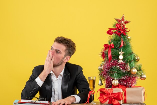 Vista frontal trabajador masculino detrás de su lugar de trabajo con regalos en escritorio amarillo trabajo de año nuevo oficina de trabajo de color de navidad