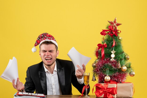 Vista frontal trabajador masculino sentado detrás de su lugar de trabajo con documentos en el escritorio amarillo oficina de trabajo año nuevo color trabajo de navidad