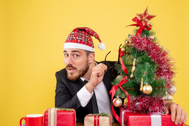 Vista frontal trabajador masculino sentado detrás de su lugar de trabajo con regalos en amarillo