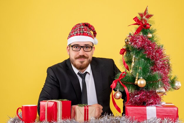 Vista frontal trabajador masculino sentado detrás de su lugar de trabajo con regalos en amarillo