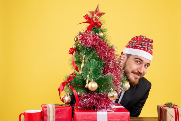 Vista frontal trabajador masculino sentado detrás de su lugar de trabajo con regalos en el escritorio amarillo trabajo oficina de navidad color regalo de año nuevo