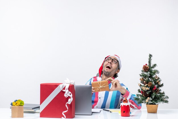 Vista frontal trabajador masculino sentado en su lugar de trabajo con paquete vacaciones emoción trabajo de oficina de navidad