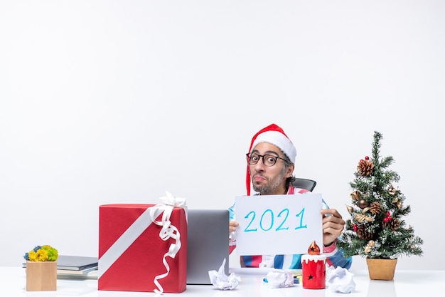 Vista frontal trabajador masculino sentado en su lugar de trabajo sosteniendo la hoja de papel con el número 2021, año nuevo concepto trabajo oficina navidad