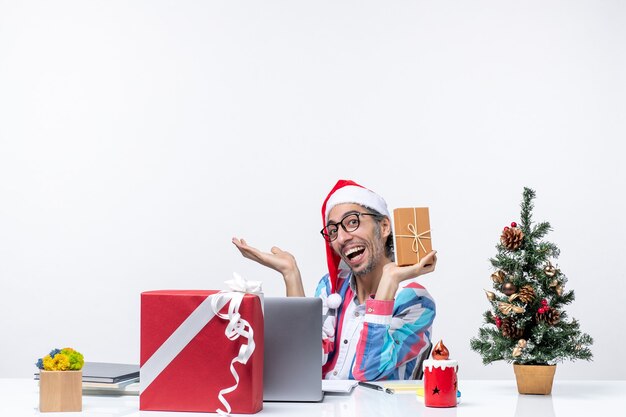 Vista frontal trabajador masculino sentado en su lugar de trabajo sosteniendo paquete humano trabajo de navidad oficina de emociones de vacaciones