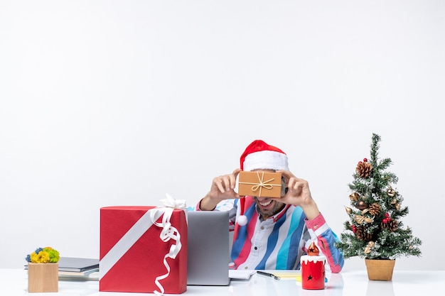 Vista frontal trabajador masculino sentado en su lugar de trabajo sosteniendo paquete vacaciones emoción oficina de trabajo de navidad