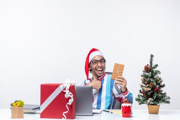 Vista frontal trabajador masculino sentado en su lugar de trabajo sosteniendo paquete vacaciones emociones trabajo oficina de negocios de navidad