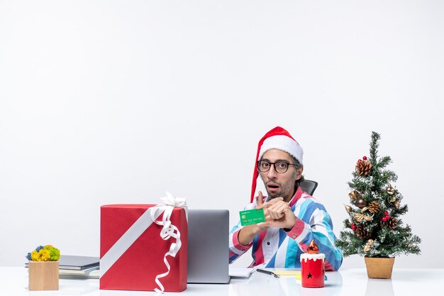 Vista frontal del trabajador masculino sentado en su lugar de trabajo sosteniendo la tarjeta bancaria verde oficina dinero de navidad trabajo emociones