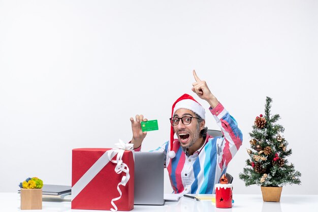 Vista frontal trabajador masculino sentado en su lugar de trabajo con tarjeta bancaria verde dinero trabajo oficina emoción de navidad
