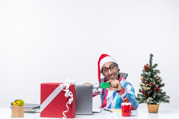 Vista frontal trabajador masculino sentado en su lugar de trabajo con tarjeta bancaria verde oficina emoción trabajo de dinero de navidad