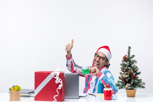 Vista frontal trabajador masculino sentado en su lugar de trabajo con tarjeta bancaria verde trabajo emoción trabajo de oficina de navidad dinero