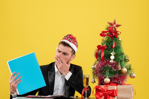 Vista frontal trabajador masculino sosteniendo documentos alrededor del pequeño árbol de Navidad y presenta en amarillo