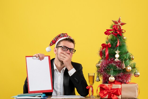 Vista frontal trabajador masculino sosteniendo nota alrededor del pequeño árbol de Navidad y presenta en amarillo