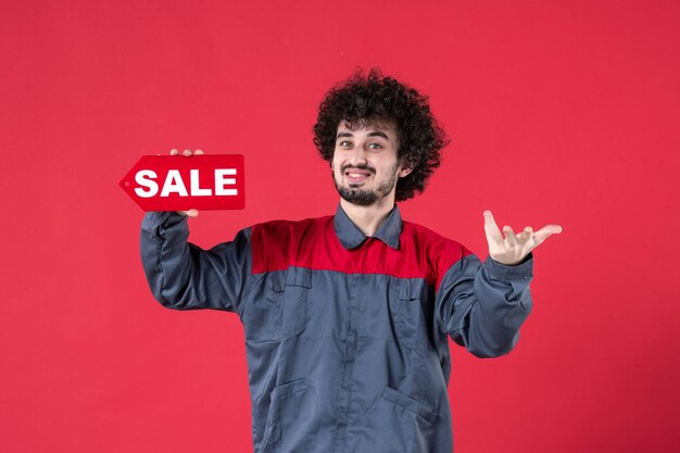 Vista frontal trabajador masculino sosteniendo la placa de venta roja sobre fondo rojo mecánico casa trabajo uniforme color de la foto del instrumento