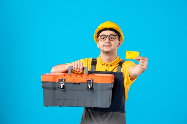 Vista frontal trabajador masculino en uniforme y caja de herramientas de casco y tarjeta bancaria en azul