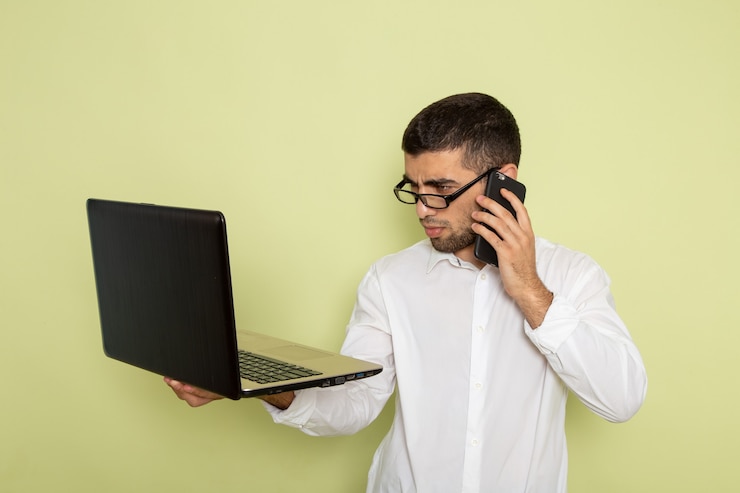 Foto gratuito vista frontal del trabajador de oficina masculino en camisa blanca hablando por teléfono y usando la computadora portátil en la pared verde