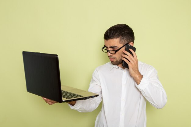 Vista frontal del trabajador de oficina masculino en camisa blanca hablando por teléfono y usando la computadora portátil en la pared verde