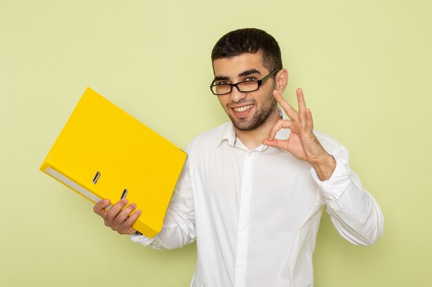 Vista frontal del trabajador de oficina masculino en camisa blanca sosteniendo un archivo amarillo y sonriendo en la pared verde claro