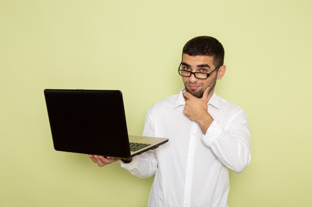 Vista frontal del trabajador de oficina masculino en camisa blanca sosteniendo y usando la computadora portátil pensando en la pared verde claro