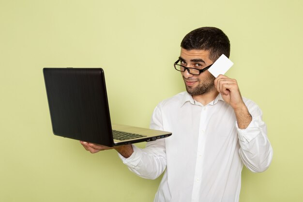 Vista frontal del trabajador de oficina masculino en camisa blanca sosteniendo y usando su computadora portátil en la pared verde claro