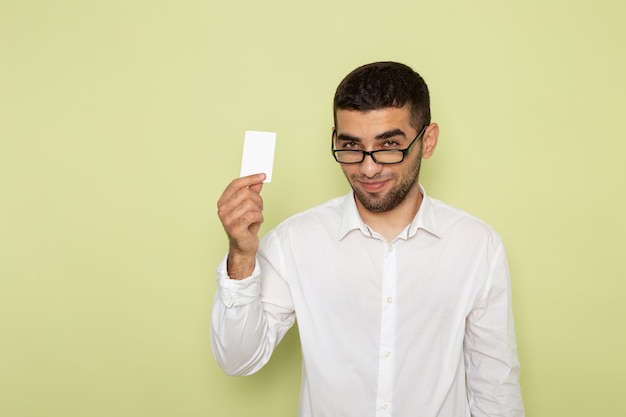 Vista frontal del trabajador de oficina masculino en camisa blanca con tarjeta con sonrisa en la pared verde claro