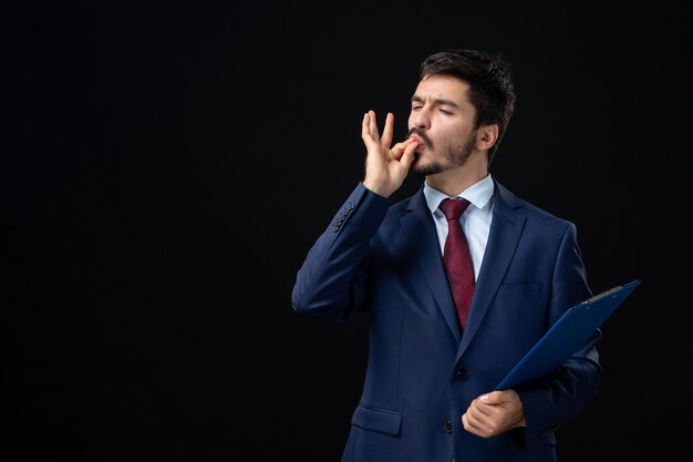 Vista frontal del trabajador de oficina masculino emocional en traje sosteniendo documentos y haciendo un gesto perfecto en la pared oscura aislada