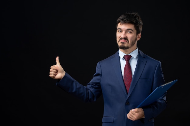 Vista frontal del trabajador de oficina masculino orgulloso y ambicioso en traje sosteniendo documentos y haciendo un gesto aceptable en la pared oscura aislada