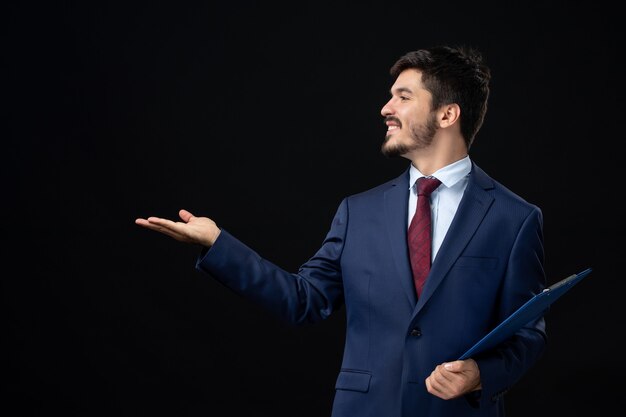 Vista frontal del trabajador de oficina masculino satisfecho en traje sosteniendo documentos y apuntando algo en el lado derecho en la pared oscura aislada