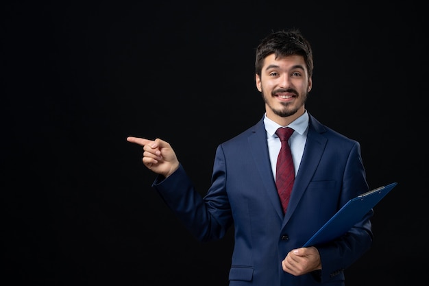 Vista frontal del trabajador de oficina masculino sonriente en traje sosteniendo documentos y apuntando algo en el lado derecho en la pared oscura aislada